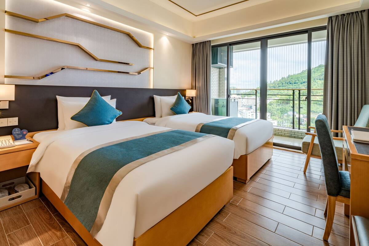 Quarto de hotel com duas camas, uma escrivaninha e vista para uma paisagem verde.