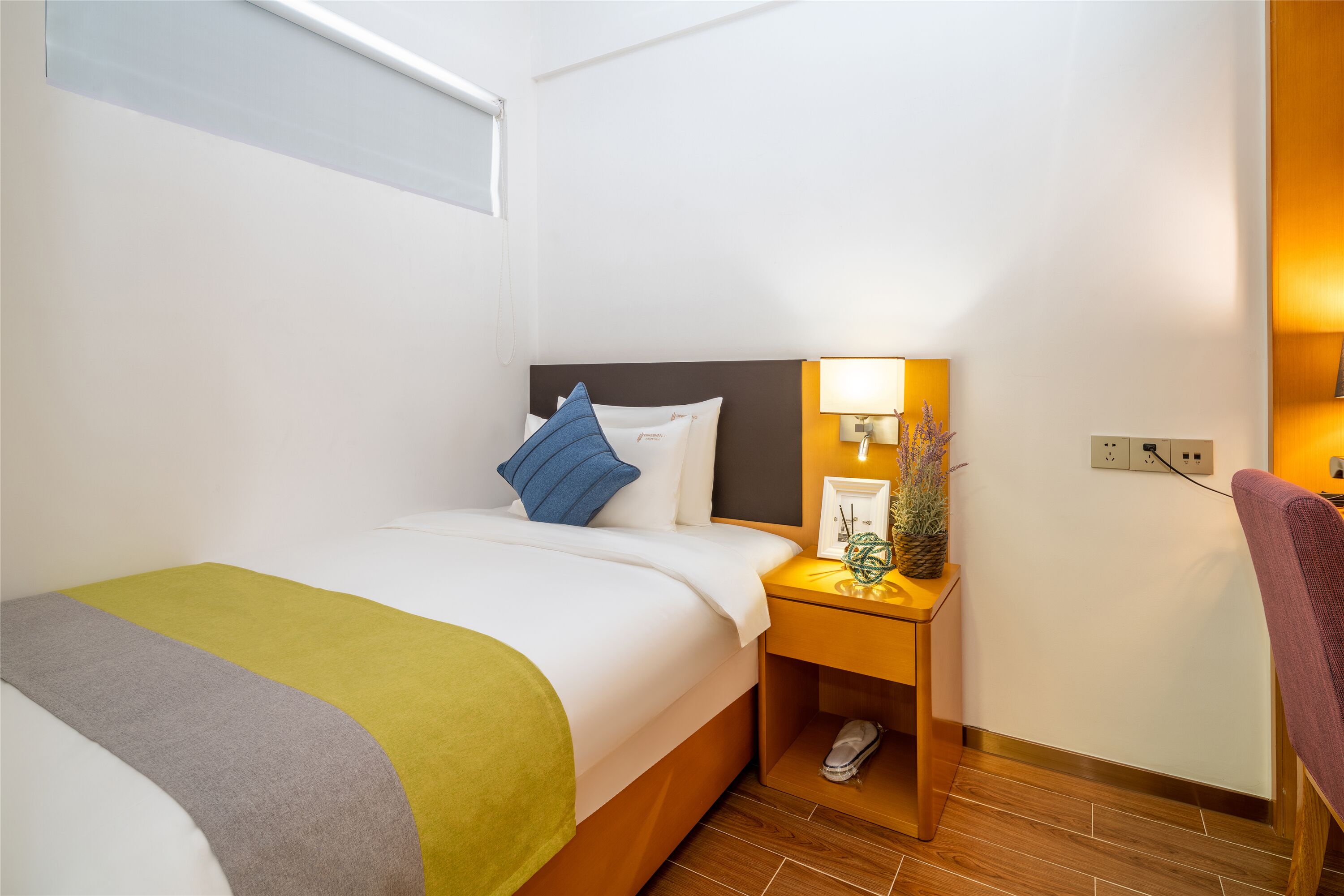 Una cama bien hecha con una manta blanca y verde, una mesita de noche con una lámpara y una planta, y un piso de madera.