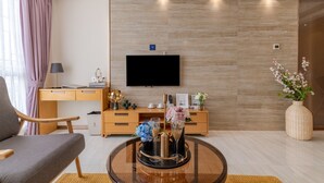 Ein modernes Wohnzimmer mit Flachbildfernseher, Holzmöbeln, einem Glastisch mit Dekoration und einem großen Fenster mit Vorhängen.