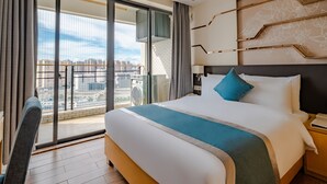 Ein Hotelzimmer mit einem großen Bett, Blick auf die Stadt durch raumhohe Fenster und einem Balkon mit Stadtblick.