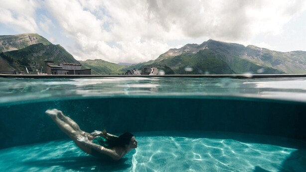 Uma pessoa nadando em uma piscina cristalina, com uma paisagem montanhosa ao fundo.