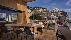 Um deck de madeira ao ar livre com mesas e cadeiras, com vista para uma aldeia montanhosa com edifícios cobertos de neve.