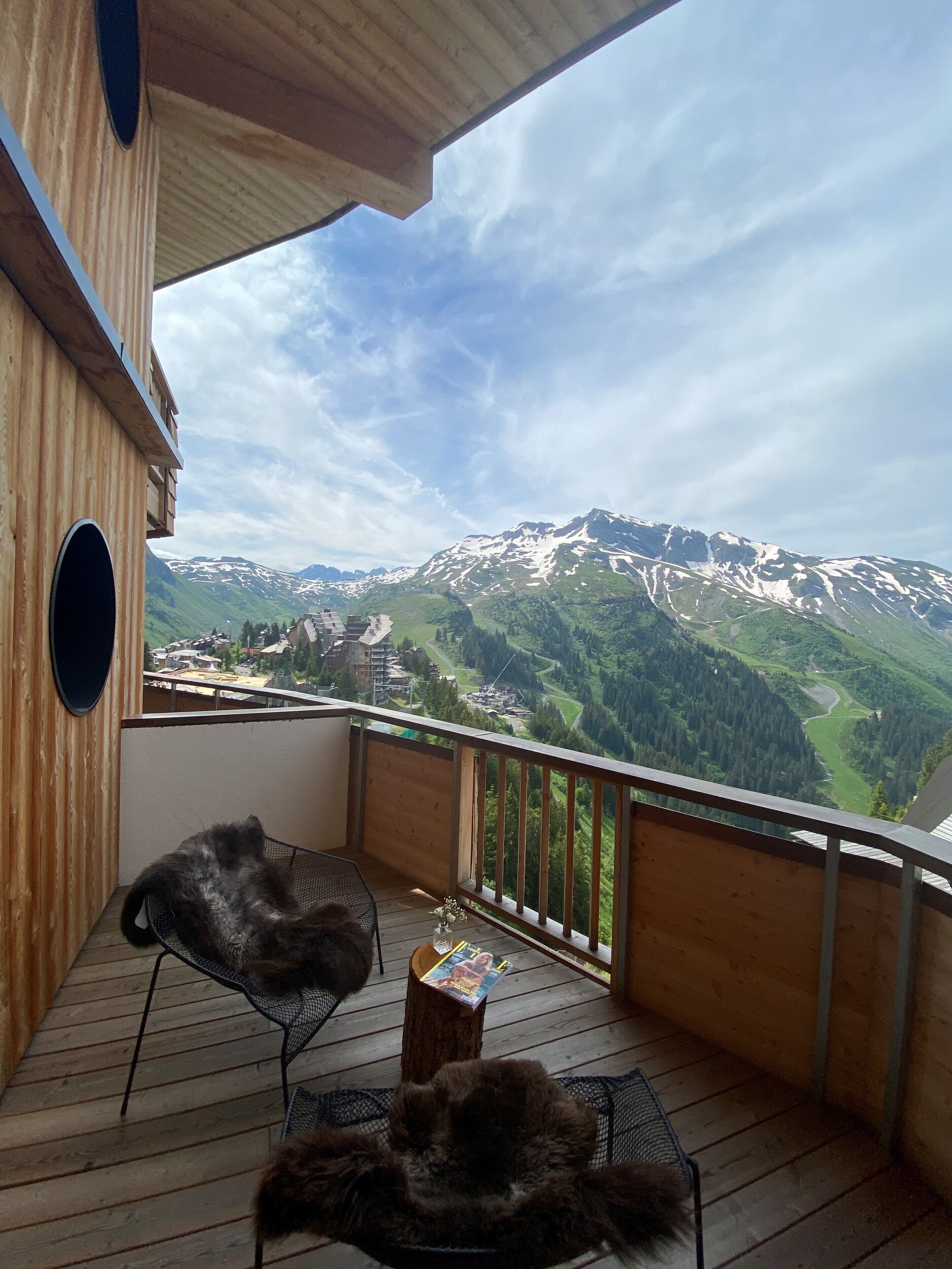 A wooden deck with two chairs and a table, overlooking a mountainous landscape with snow-capped peaks and a valley below.