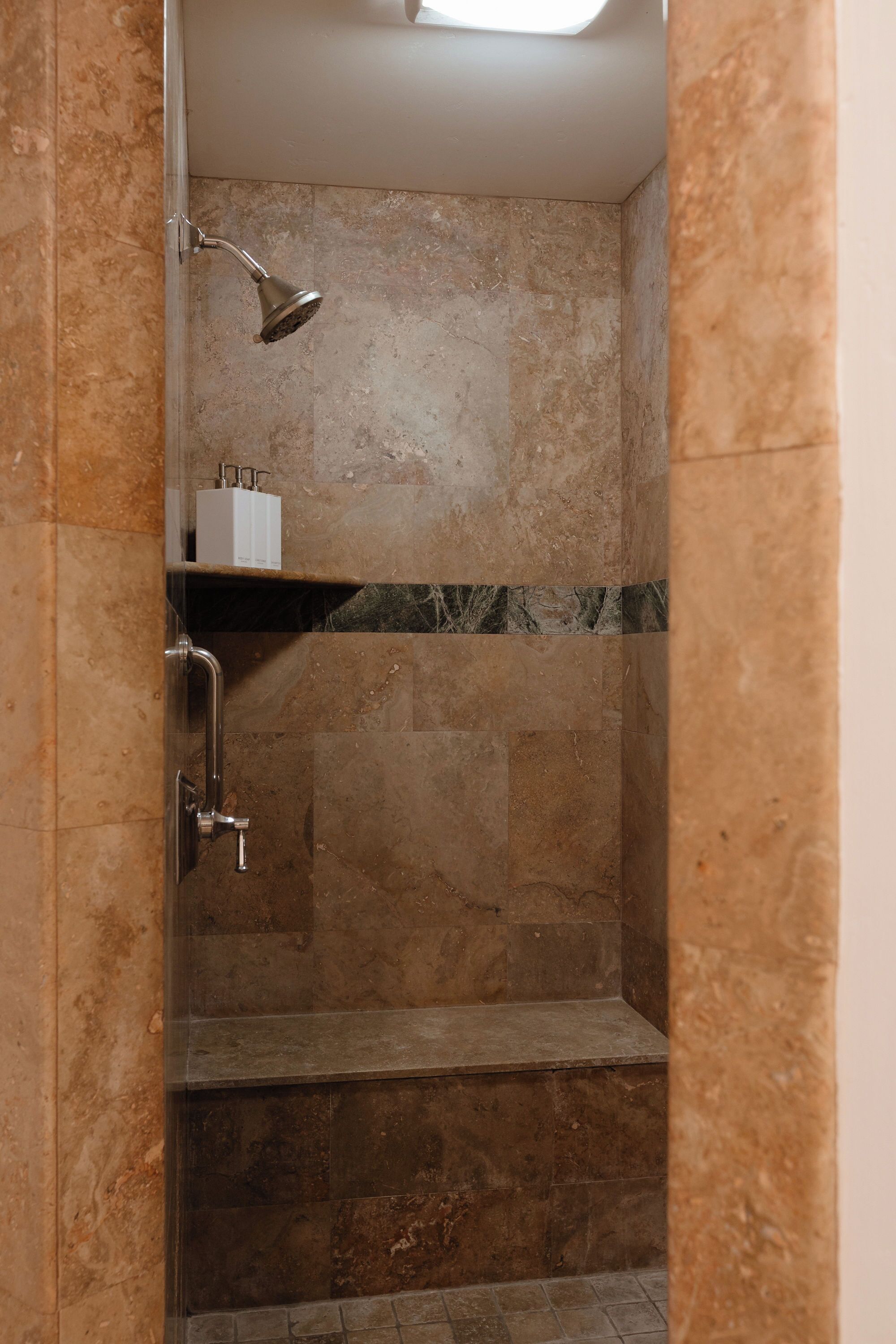 A shower with a handheld showerhead, a shelf with toiletries, and a marble-tiled wall.
