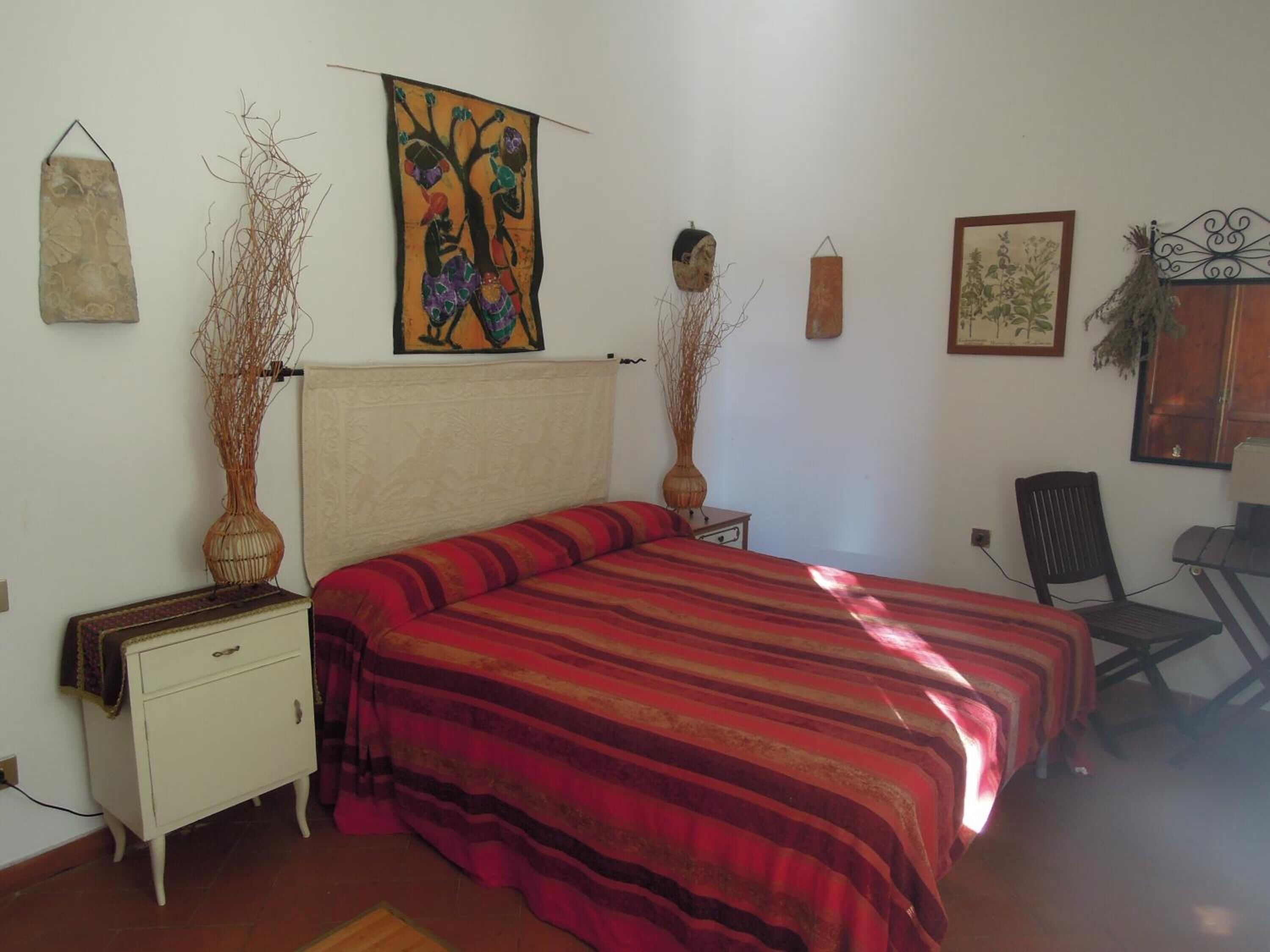 A bedroom with a bed, a nightstand, a chair, and wall decorations including a painting and two vases.
