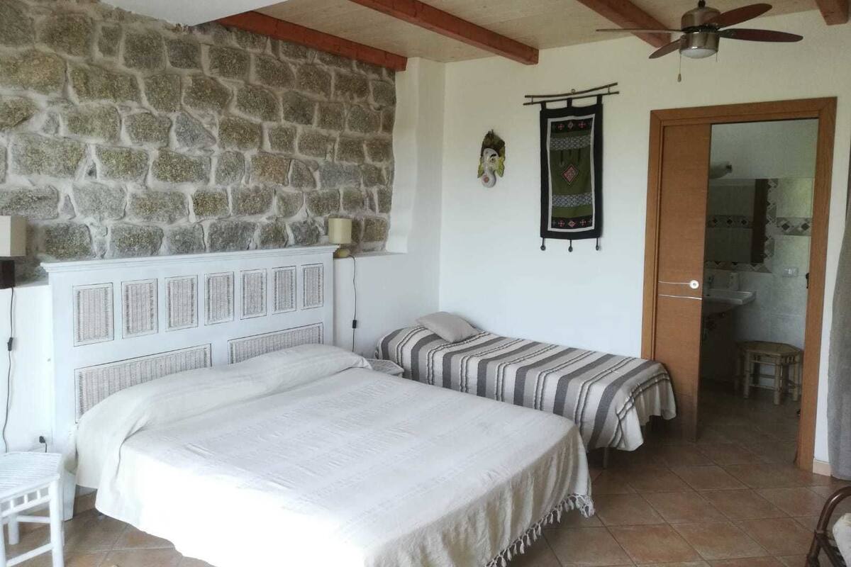 Une chambre avec un mur en pierre, un lit avec une tête de lit, une table de chevet, une lampe et une porte donnant sur une salle de bain.