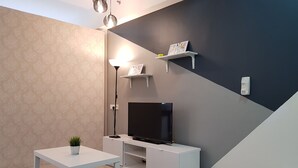 Una sala de estar moderna con un televisor de pantalla plana sobre un soporte blanco, una pequeña mesa blanca con una planta, un tapete a rayas y una lámpara.