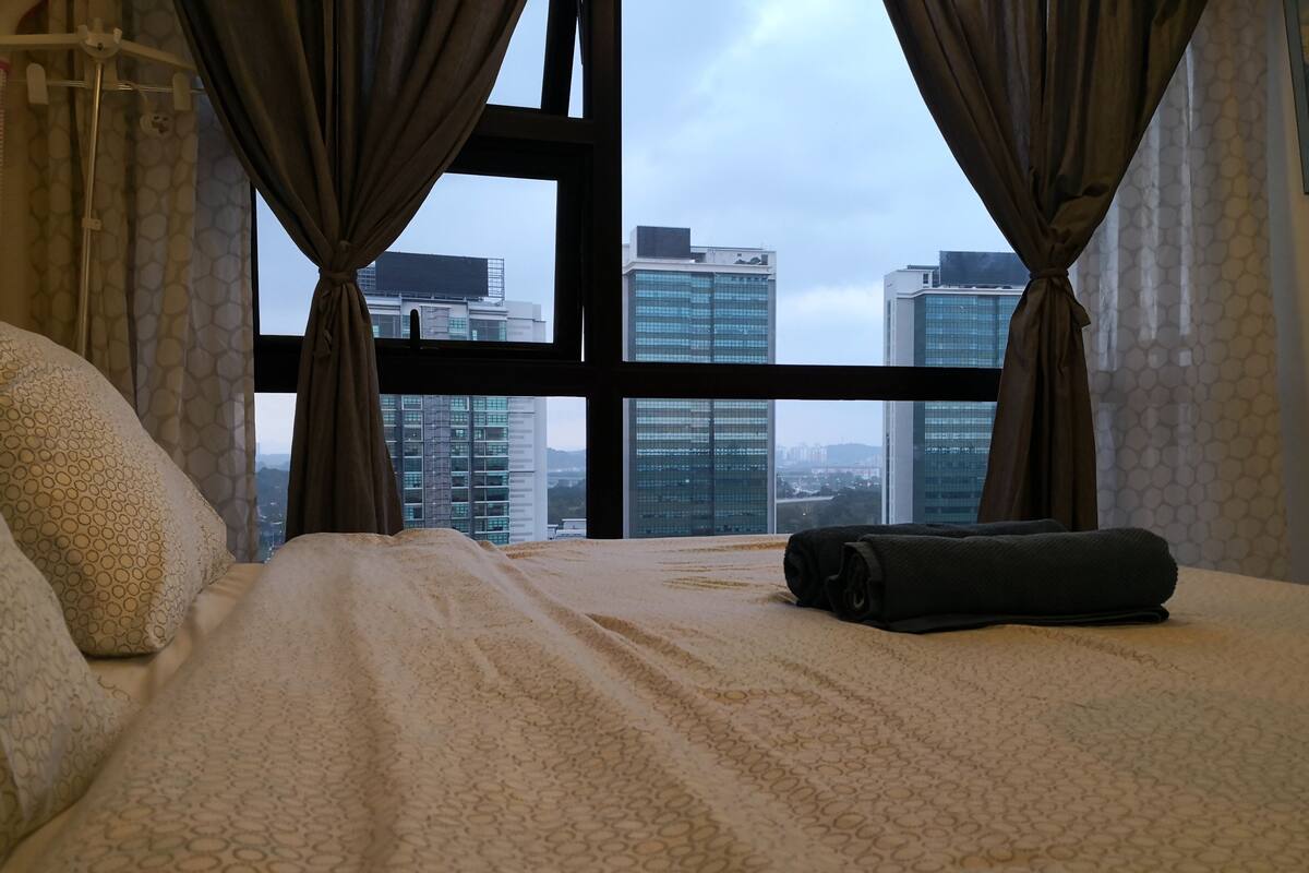 A hotel room with a large bed, a view of tall buildings, and a window with curtains.