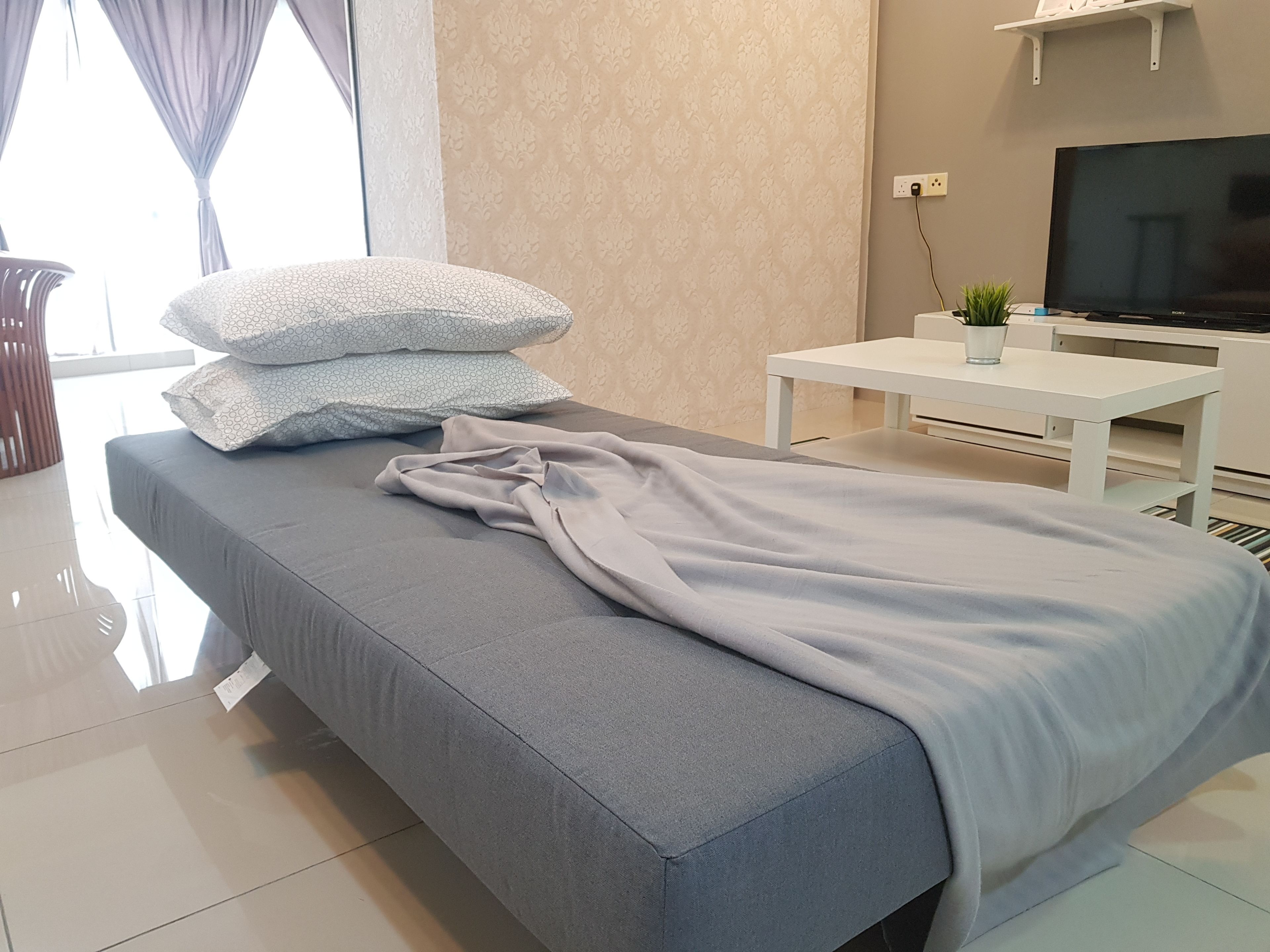 A modern bedroom with a grey bed, a white bedside table with a plant, and a TV on a stand.