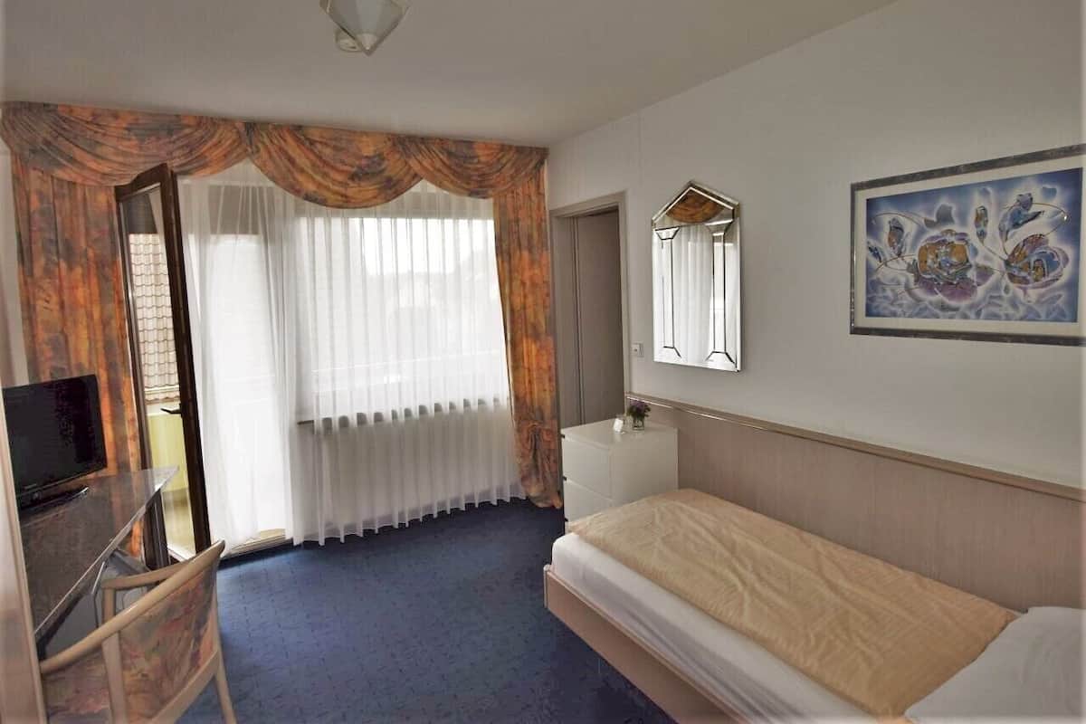 Ett hotellrum med en säng, ett skrivbord med en stol, en tv, en byrå och ett fönster med gardiner.