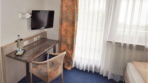 A hotel room with a bed, a desk with a chair, a TV mounted on the wall, and a window with sheer curtains. - Hotel Alena (Filderstadt)