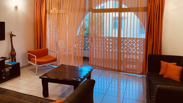 A living room with a sofa, a chair, a coffee table, and a TV. There is a window with curtains and a view of a building outside. - Barizi Holiday Apartments (Mtwapa)