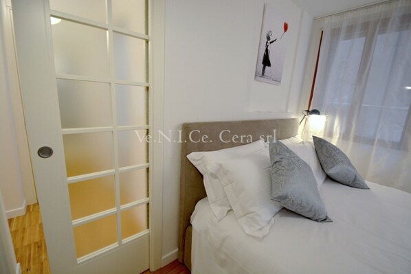 Une chambre à coucher avec un lit, des oreillers, une table de chevet, une lampe et une fenêtre avec des rideaux.