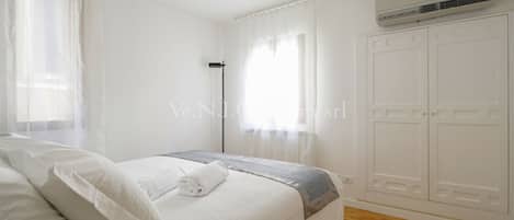 A bedroom with a bed, white curtains, an air conditioning unit, and a wardrobe.