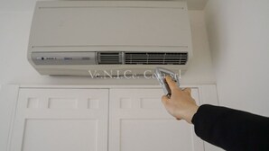 Mão segurando um controle remoto próximo a um aparelho de ar condicionado branco fixado na parede, acima de uma porta branca.