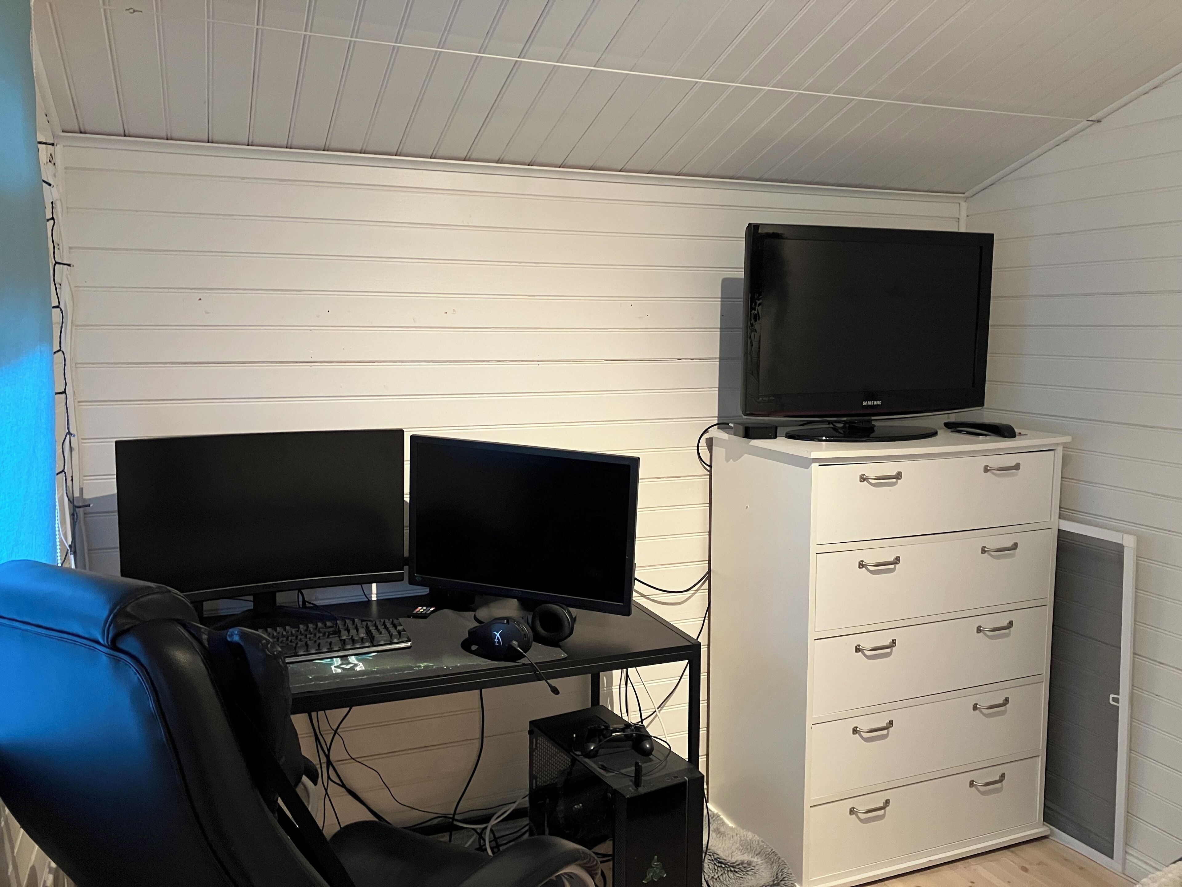 Een kamer met een bureau, twee monitoren, een toetsenbord, een muis en een gaming stoel. Er staat een televisie op een kast en een klein bed met een deken.