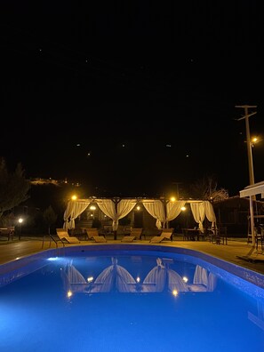 Zona de piscina nocturna con sillones de descanso y toldos, rodeada de árboles y edificios.