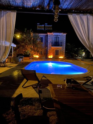 Piscina con una casa de fachada de piedra al fondo, rodeada de mobiliario y luces para exteriores.