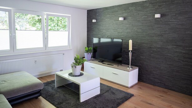 Un salon moderne doté d’un téléviseur à écran plat, de meubles blancs, d’un mur d’accent en briques sombres et de grandes fenêtres.