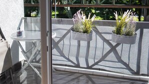 Un balcon avec une table et des chaises en verre, trois plantes en pot et une vue sur des arbres et un pont.
