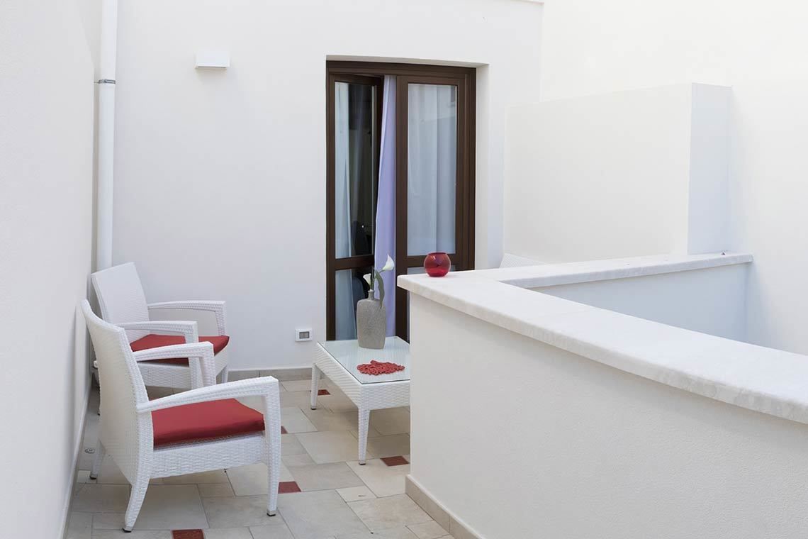 Un pequeño balcón con mobiliario de mimbre blanco y un cojín rojo, un lavamanos blanco y una ventana con cortinas.