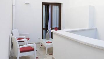 Un pequeño balcón con mobiliario de mimbre blanco y un cojín rojo, un lavamanos blanco y una ventana con cortinas.