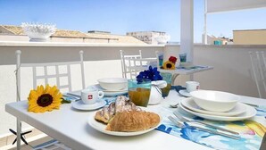 Une table dressée avec des aliments pour le petit-déjeuner, notamment des croissants et une décoration en forme de tournesol, sur un balcon d’où l’on a une vue dégagée sur les bâtiments en arrière-plan.