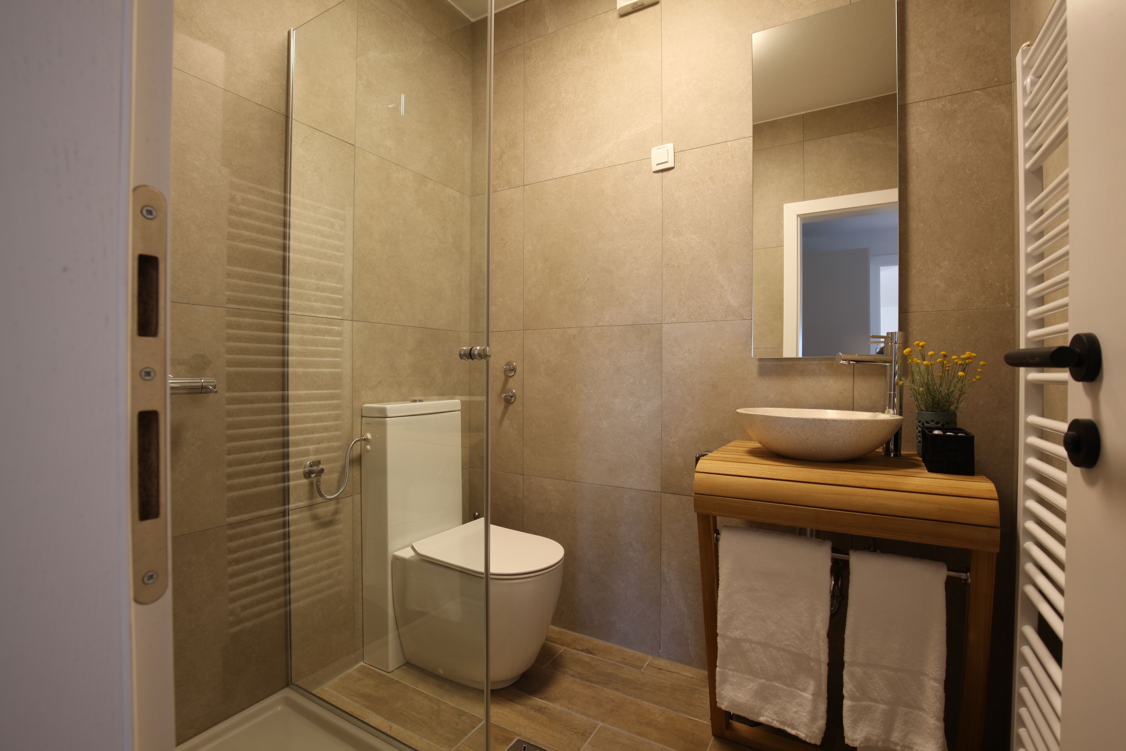 Un baño moderno con cabina de ducha de vidrio, un inodoro blanco, un tocador de madera con lavamanos y un espejo sobre el tocador.