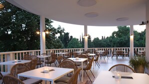 An outdoor dining area with white tables and wicker chairs, surrounded by trees. - Hotel Ortus Residence (Ankaran)