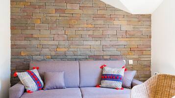 Una sala de estar con pared de piedra, un sofá morado, dos cojines estampados, una mesa de centro de madera y una silla de mimbre.