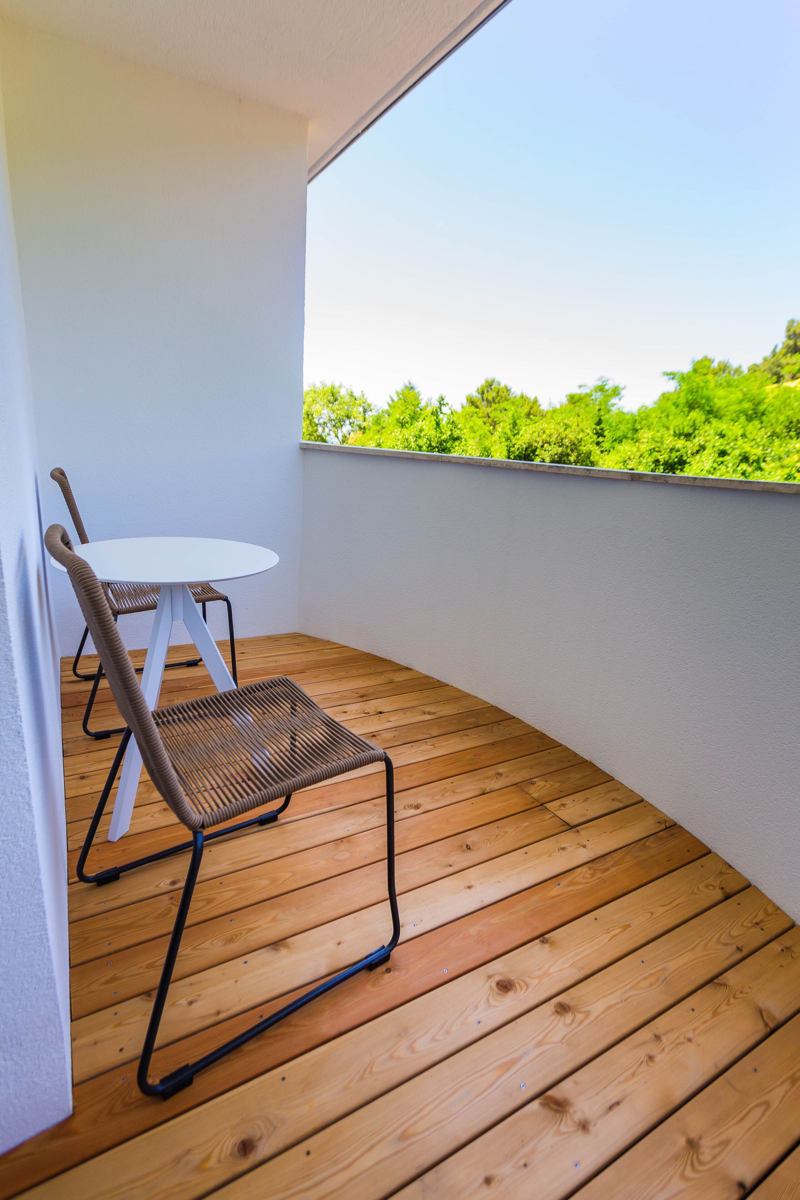 Un balcón con suelo de madera, una mesa blanca y dos sillas de mimbre.