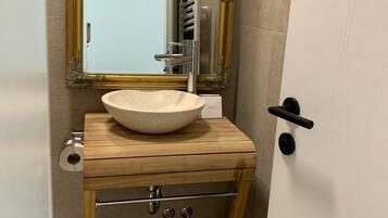 Een badkamer met een houten wastafelmeubel, een grote spiegel met een sierlijk gouden frame, een witte wasbak en een handdoekhouder.