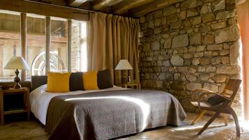 A bedroom with a stone wall, a bed with a patterned bedspread, two bedside lamps, a wooden chair, and a window with curtains.