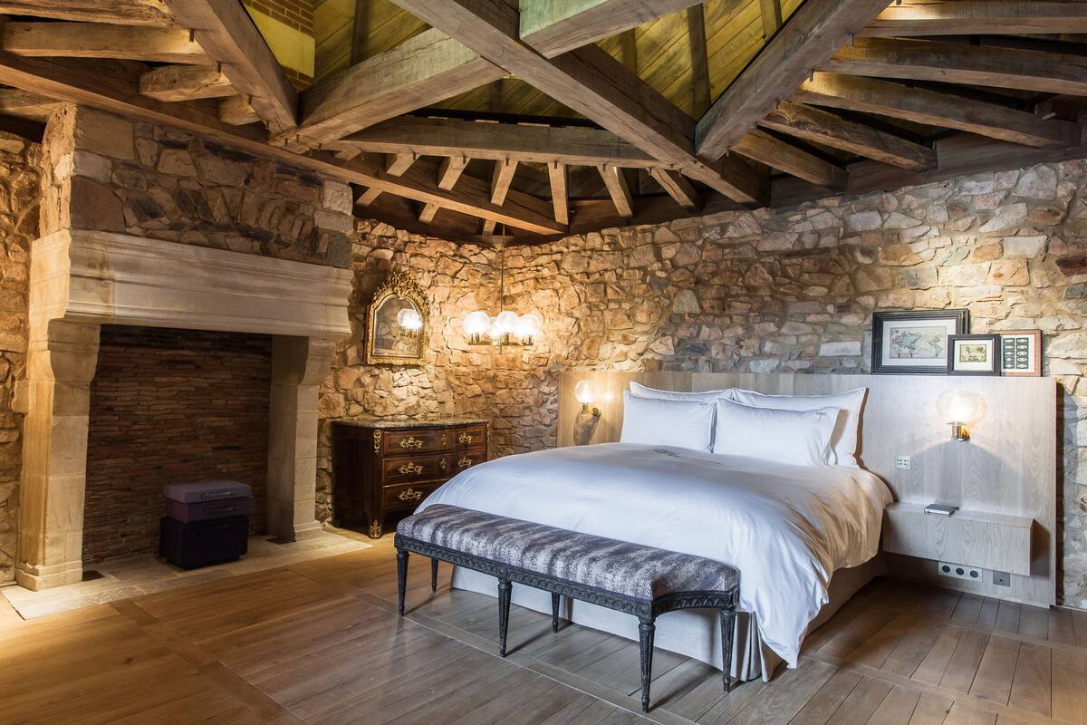 Une chambre avec un mur en pierre, des poutres apparentes, une cheminée, un lit, des tables de chevet et des lampes fixées au mur.