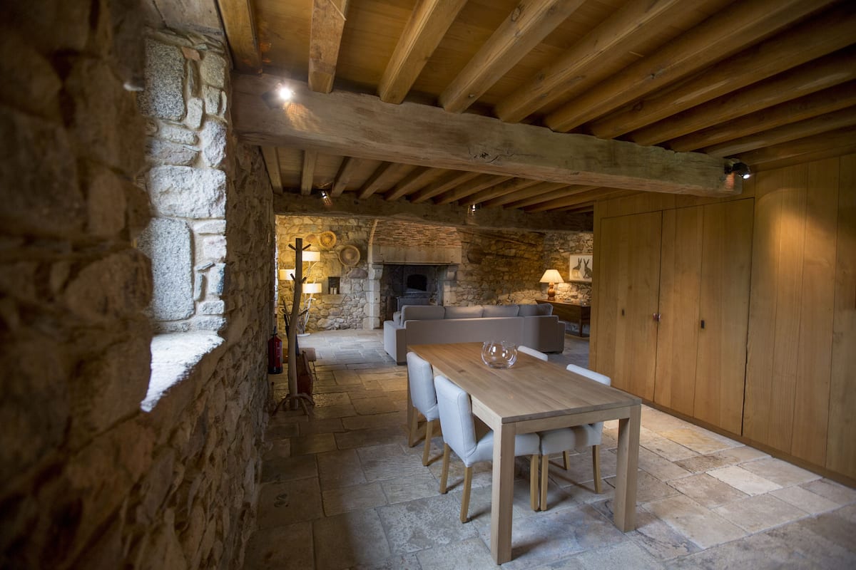 Une pièce rustique aux murs de pierre, avec un plafond en bois, une cheminée en pierre, une table à manger en bois entourée de chaises et un canapé.
