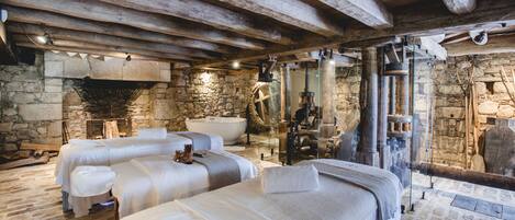 A rustic room with stone walls, wooden beams, and a stone fireplace. There are three beds with white linens and a large stone bathtub.