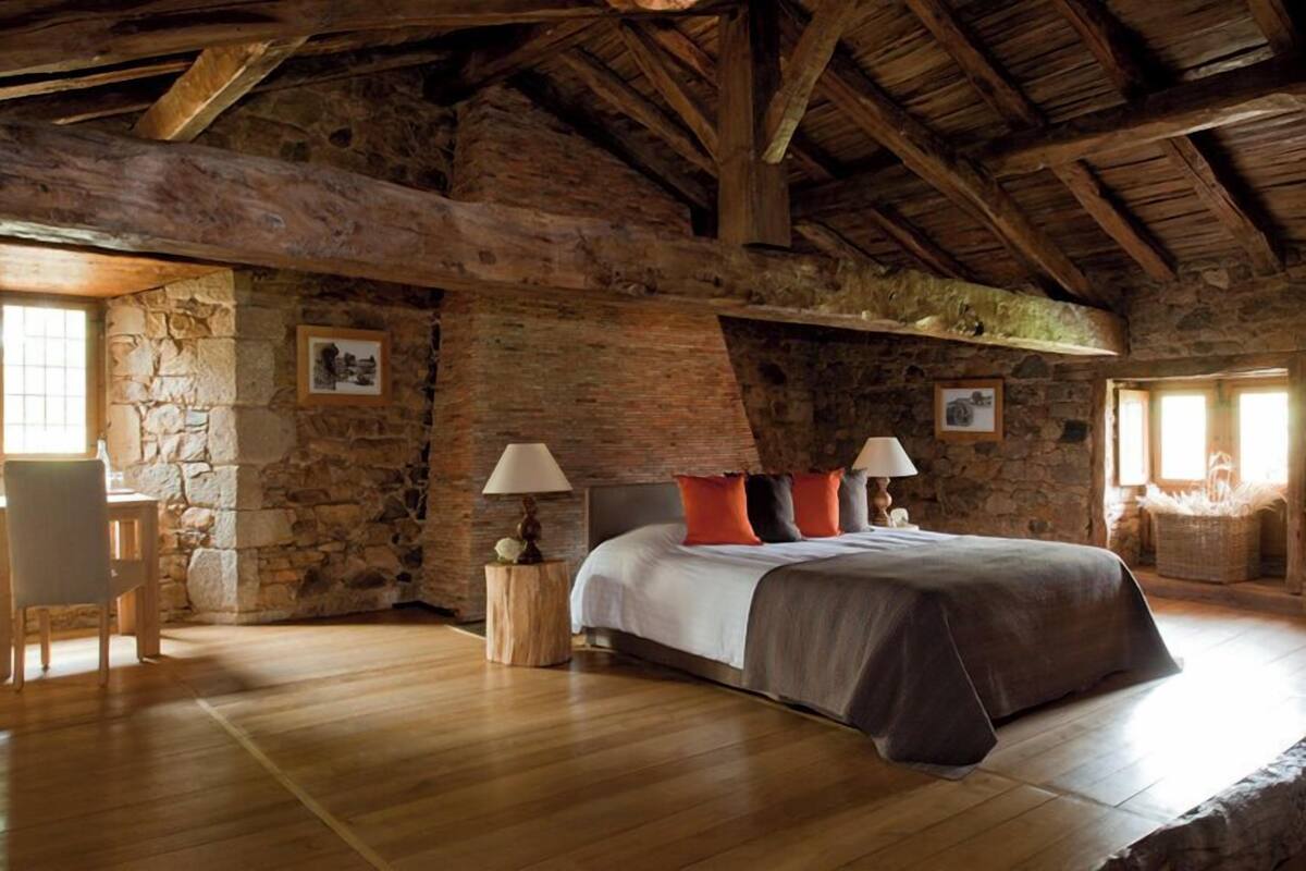 Une chambre à coucher rustique avec un mur en pierre, des poutres en bois, un grand lit avec des oreillers, deux tables de chevet avec des lampes et un fauteuil.