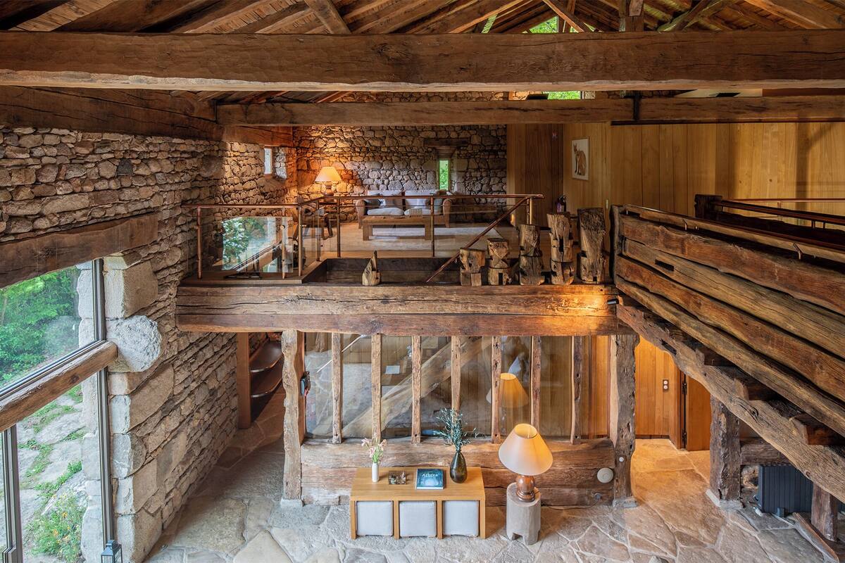 L’intérieur d’un chalet rustique, avec un mur en pierre, des poutres en bois, un escalier en bois et un coin salon confortable agrémenté de coussins et d’une lampe.