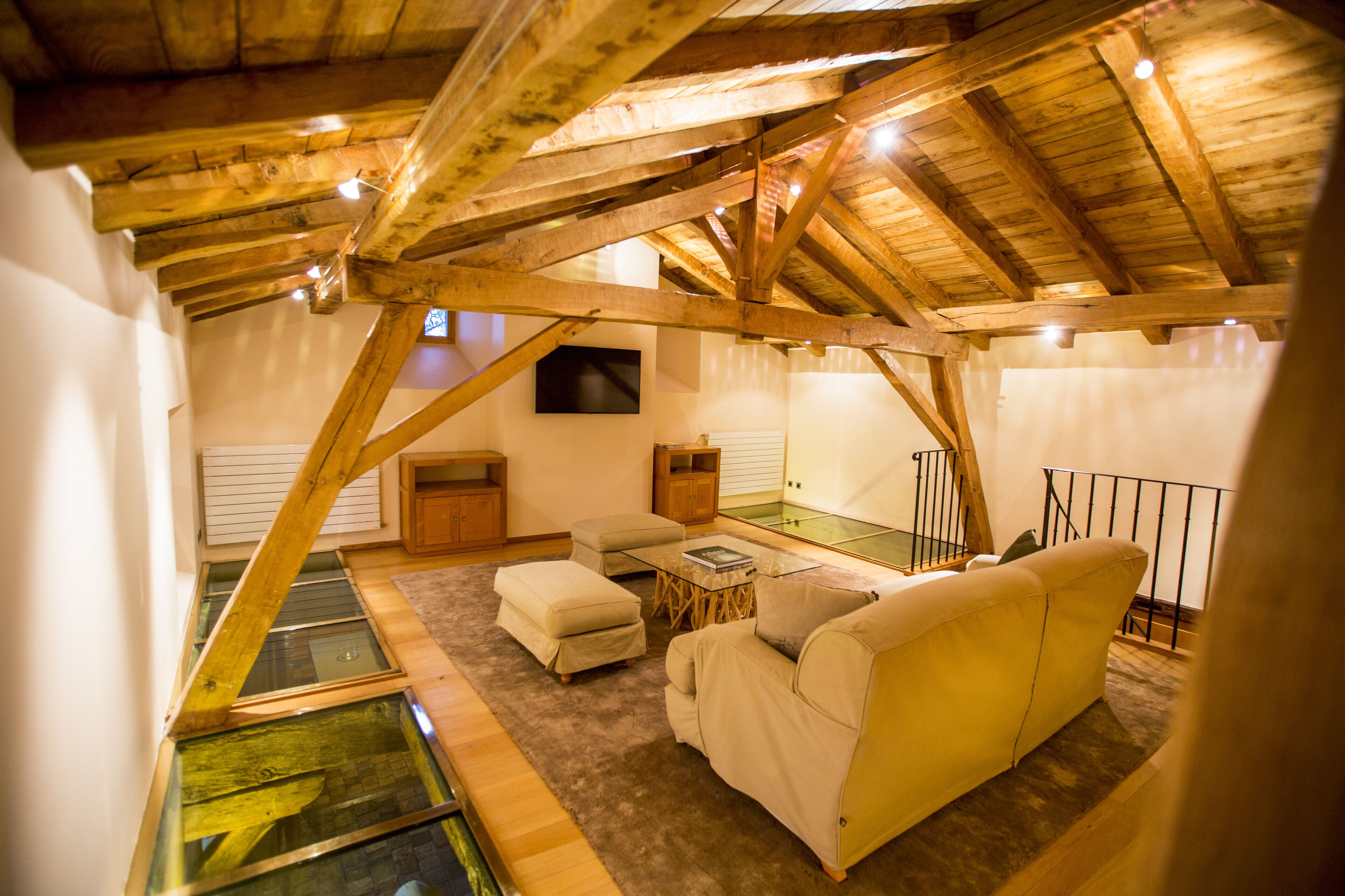A cozy loft bedroom with a sloped ceiling, wooden beams, a glass floor, a sofa, a small table, and a TV mounted on the wall.