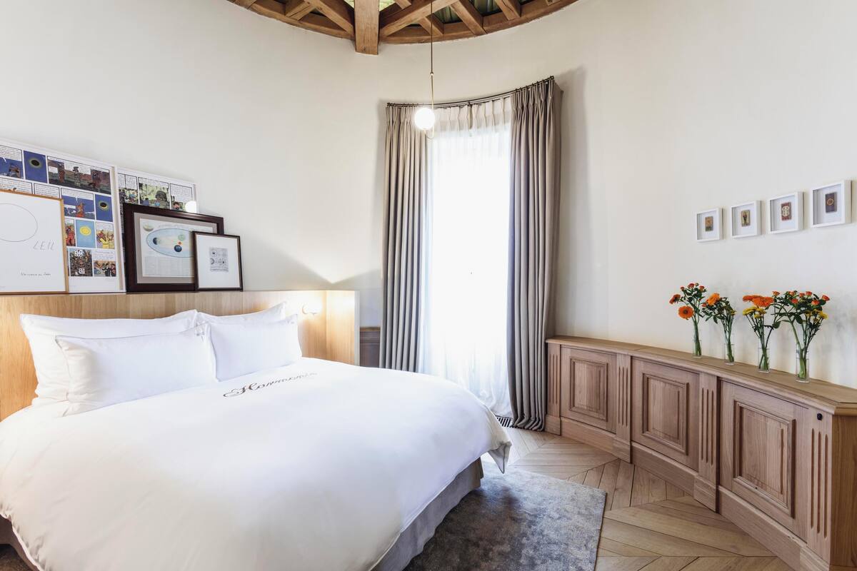 Une chambre à coucher bien aménagée, avec un grand lit, une tête de lit en bois, une table de chevet avec une lampe, une fenêtre avec des rideaux et un cadre photo fixé au mur.