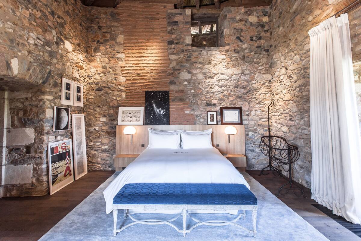 Une chambre avec un mur en pierre, un grand lit, deux tables de chevet, des tableaux encadrés et une lampe sur pied.