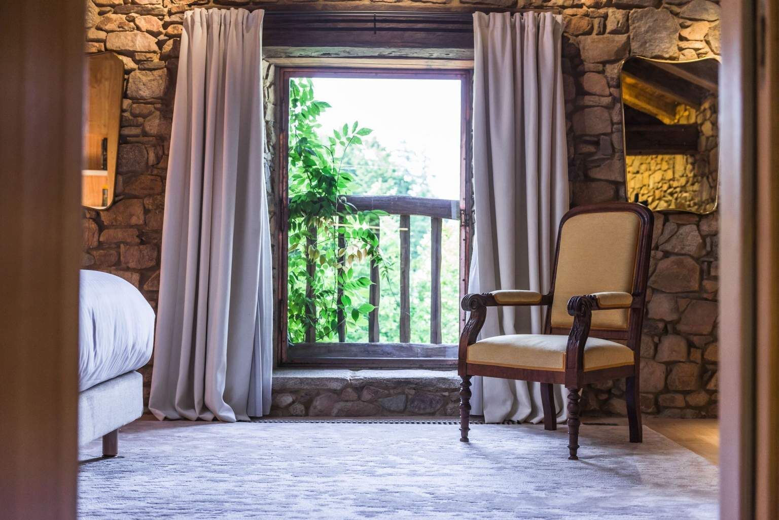 Una habitación con pared de piedra, una ventana con cortinas, una silla de madera y una cama.