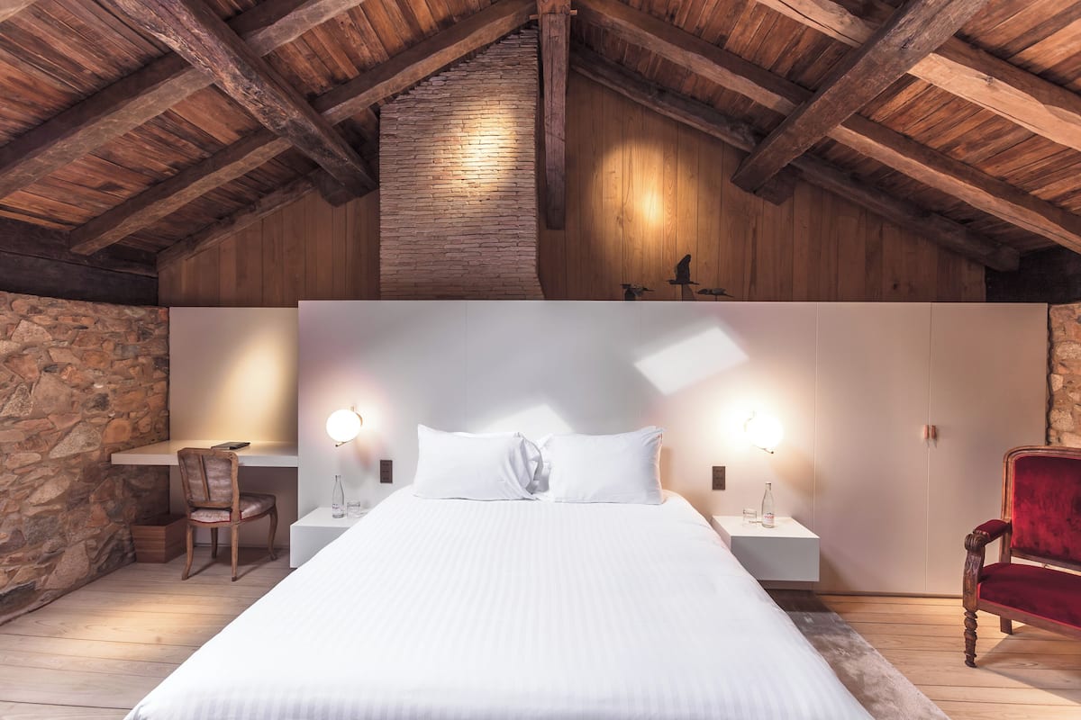 Une chambre avec un mur en pierre, des poutres en bois au plafond, un lit avec du linge de maison blanc, une chaise rouge et des tables de chevet avec des lampes.