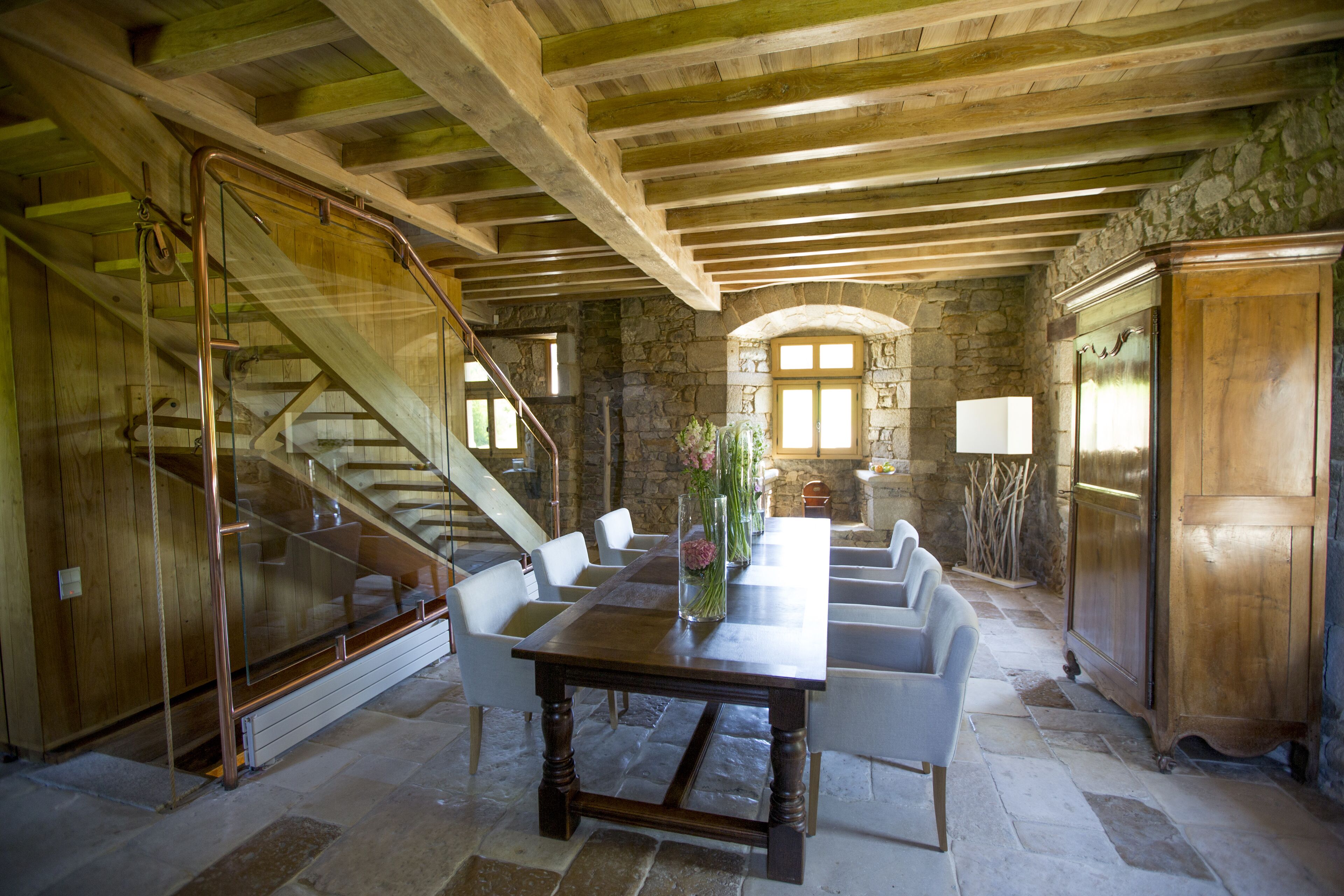 Una habitación amplia con una escalera de madera, paredes de piedra y una gran mesa de comedor con sillas.