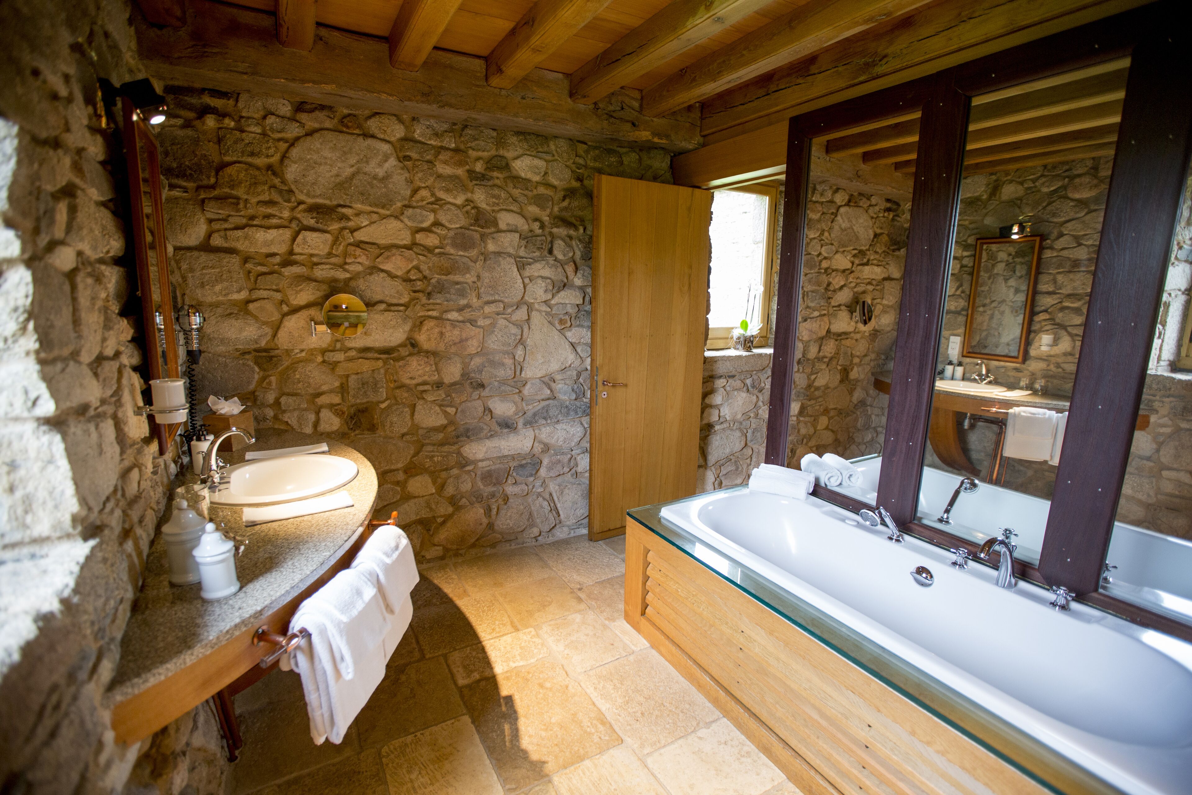 A stone-walled bathroom with a wooden vanity, a large bathtub, and a mirror with a wooden frame.