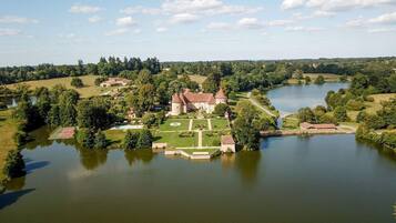 A large estate with a central building surrounded by a moat, a tennis court, and a lake.