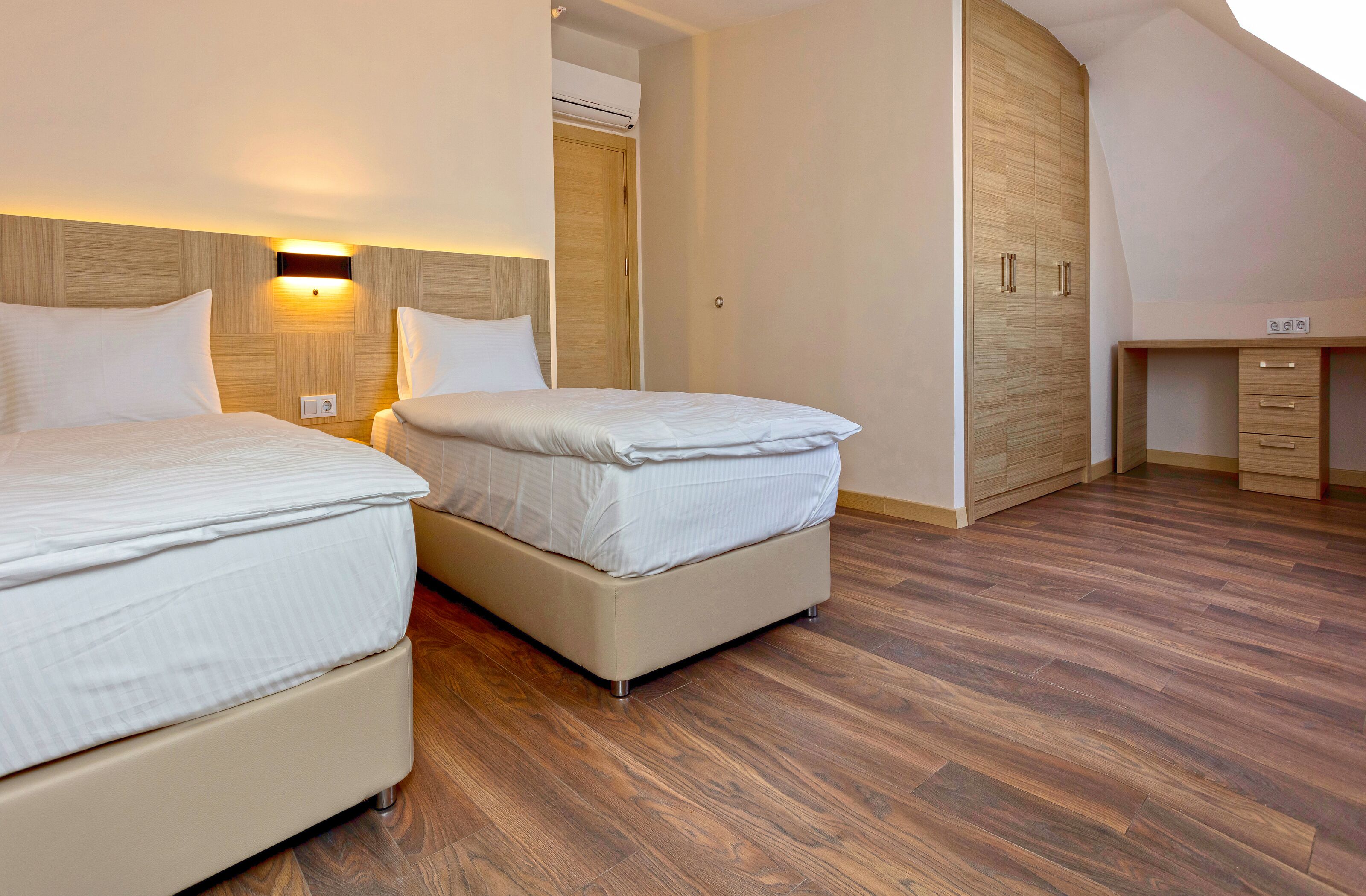 A modern hotel room with two beds, wooden flooring, a built-in wardrobe, and a small desk area.
