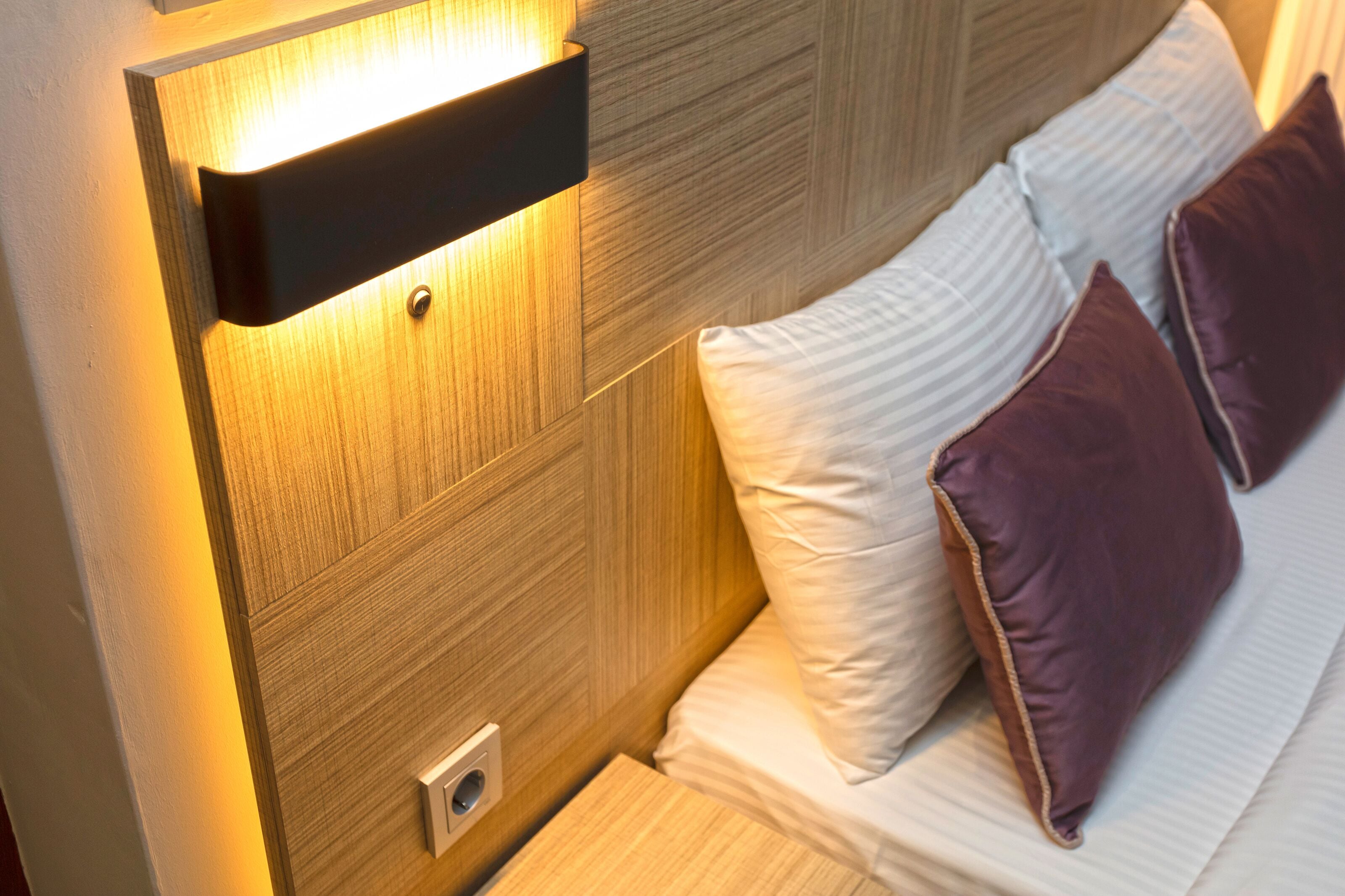 Cabecera de madera con una lámpara empotrada, colchón blanco con dos almohadas y una mesita de noche pequeña.