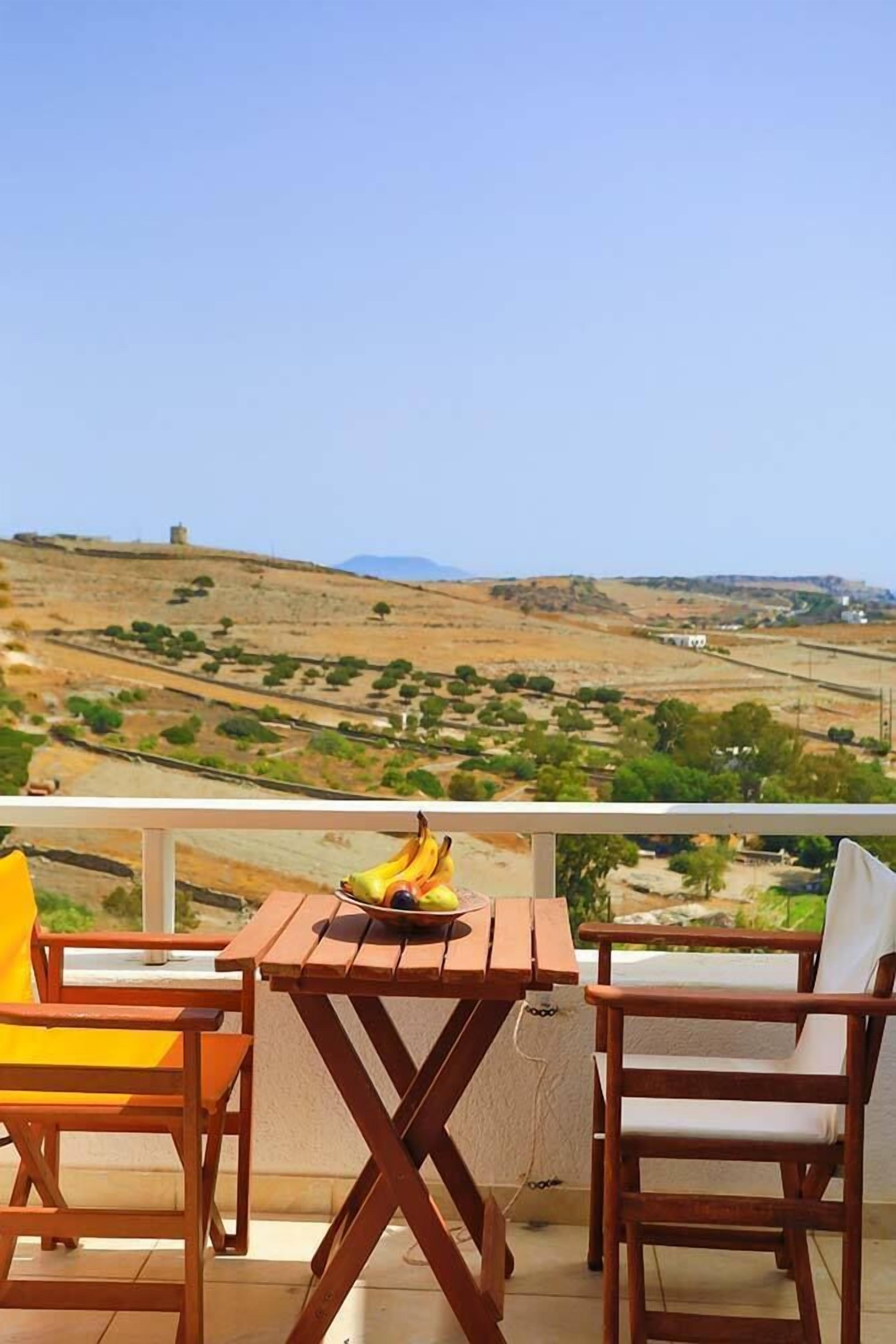 A wooden table and chairs on a balcony overlooking a hilly landscape.