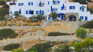 Een gebouw aan de kust met witte muren en blauwe details, gelegen op een heuvel met rotsachtig terrein en schaarse begroeiing.